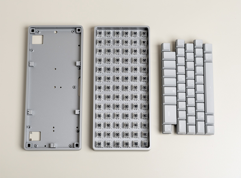 Three keyboard parts laid out on a desk: a barebones hotswap board, a tray of switches, and a PBT keycap set