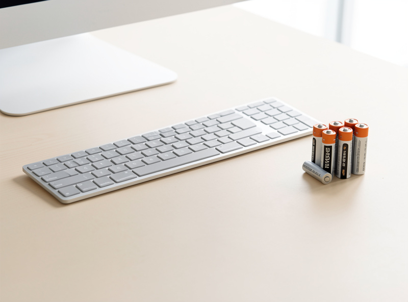 A slim low-profile wireless keyboard beside a set of rechargeable AA batteries
